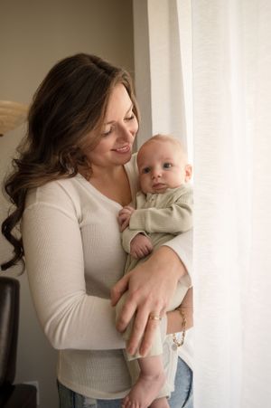 Mother in cream sweater lovingly holds small baby near window in tender moment.
