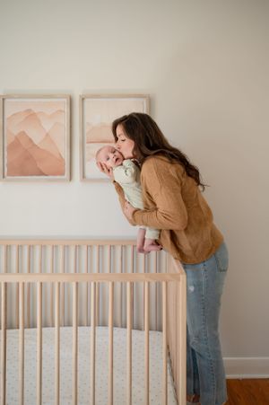 Tender nursery moment with caregiver and infant by wooden crib under pink mountain artwork.