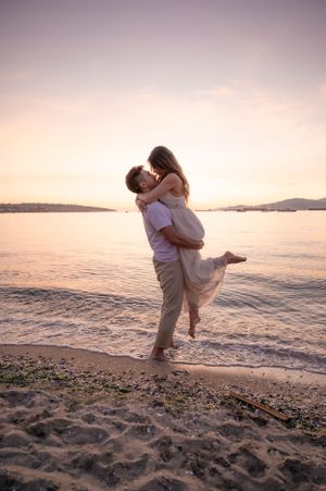 Romantic couple embraces on beach during sunset with water and mountains in background.
