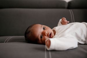A baby in white clothing lies on a dark grey leather couch looking at the camera.
