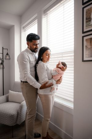 A couple stands by a bright window in their home, holding their newborn baby.