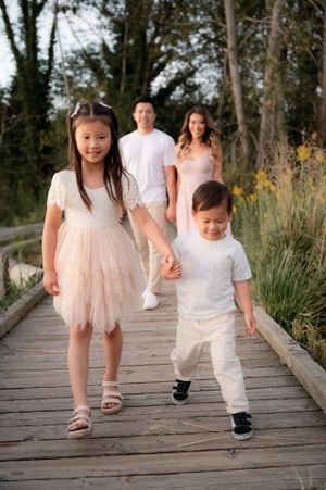 Family walks together on wooden boardwalk surrounded by tall grass and trees.