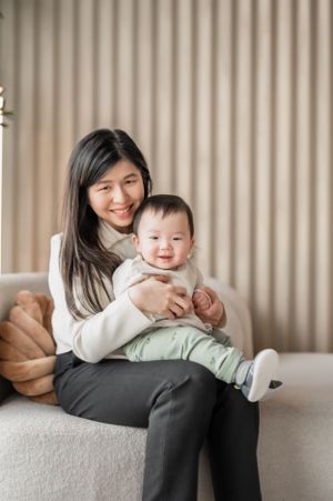 A parent and baby sit together on a light gray couch, sharing a tender moment against a beige curtain backdrop.