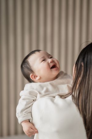 A joyful baby in a white sweater laughs while being held against a beige curtained background.