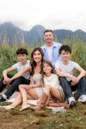 A family group sits together in casual white clothing on a grassy hill with mountain peaks visible in the background.