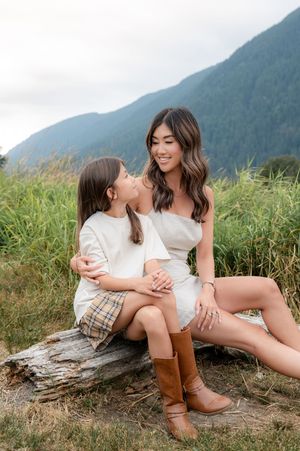 Two people in white outfits sitting on a log against a mountainous backdrop with tall grass and brown boots.