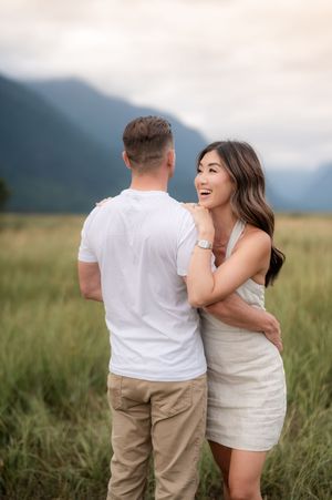 A couple shares a romantic moment while walking through a scenic grassy field with mountains in the background.