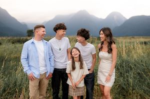 Happy group of people laugh together while standing in a grassy field with mountains in the background at sunset.