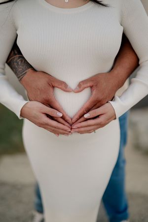 A progressive sequence of hands making a heart shape across a pregnant belly in cream colored sweater against blue jeans.