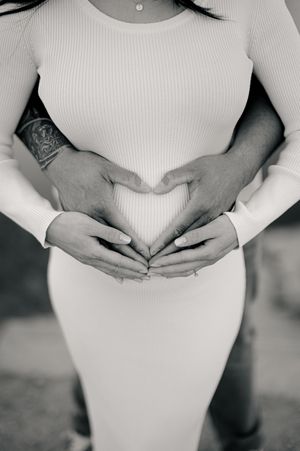 Black and white maternity photo series shows hands forming heart shapes over a pregnant belly in white clothing.