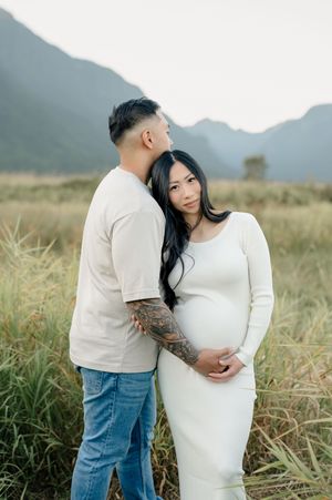 Loving couple in casual attire embraces during maternity photoshoot in scenic mountain field at golden hour.