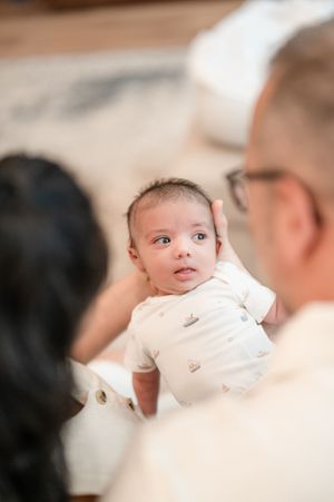 A baby in white clothing looks to the side while being held by someone wearing light colored clothing.