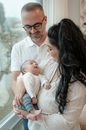 A small baby in white clothing being held tenderly by two adults near a bright window.