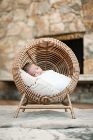 A sleeping baby wrapped in white swaddle rests in a circular rattan chair against a stone wall backdrop.