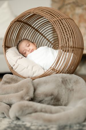 A sleeping baby wrapped in white swaddle nestled in a curved woven basket with soft blankets.