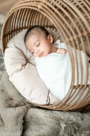 Sleeping baby wrapped in white blanket lies peacefully in curved wooden basket with cozy neutral tones.