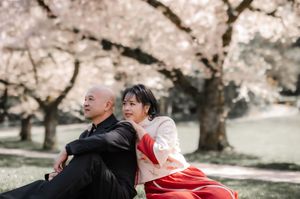 Two people sit back to back on a bench underneath blooming cherry blossom trees on a spring day.