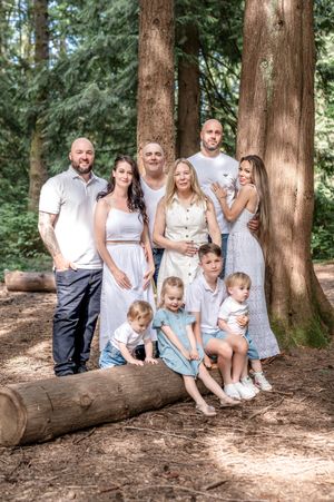 A family wearing coordinated white and grey outfits poses casually on a fallen log in a forest setting.