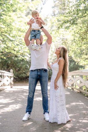 A family shares a joyful moment together on a wooden bridge path surrounded by lush green trees on a sunny day.