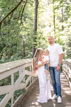A couple in white outfits shares romantic moments on a wooden bridge pathway surrounded by lush forest scenery.