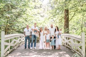 A group of people in light colored clothing stand together on a white wooden bridge surrounded by lush green trees.