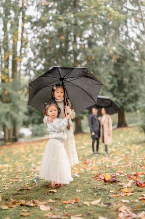 People with black umbrellas stand in a park during autumn with fallen leaves scattered on the ground.