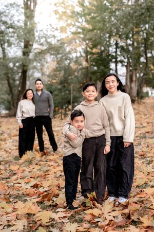 A family in coordinated beige and black outfits poses together among fallen autumn leaves in an outdoor setting.