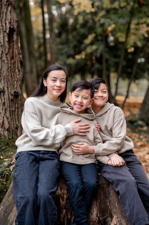 Three siblings in matching beige sweaters and dark pants sit together on a fallen tree trunk in an autumn forest.