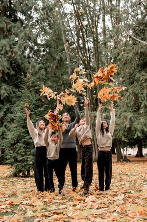 A group in black and beige outfits throws autumn leaves in the air while celebrating in a wooded outdoor setting.