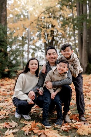 Family sits together on autumn ground covered in fallen orange leaves in matching beige sweaters.