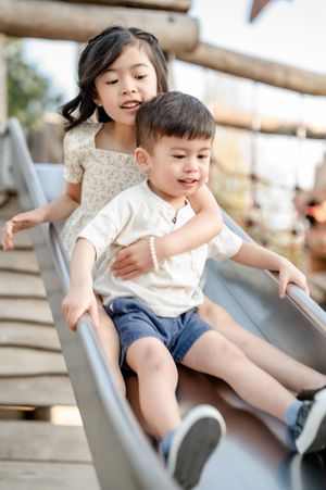 Two young Asian children joyfully sliding down a playground slide together, with the older girl hugging the younger boy.
