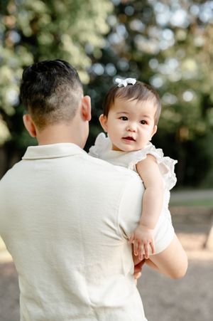 A father holds his baby girl in a white dress outdoors, she looks over his shoulder in a sunny park.