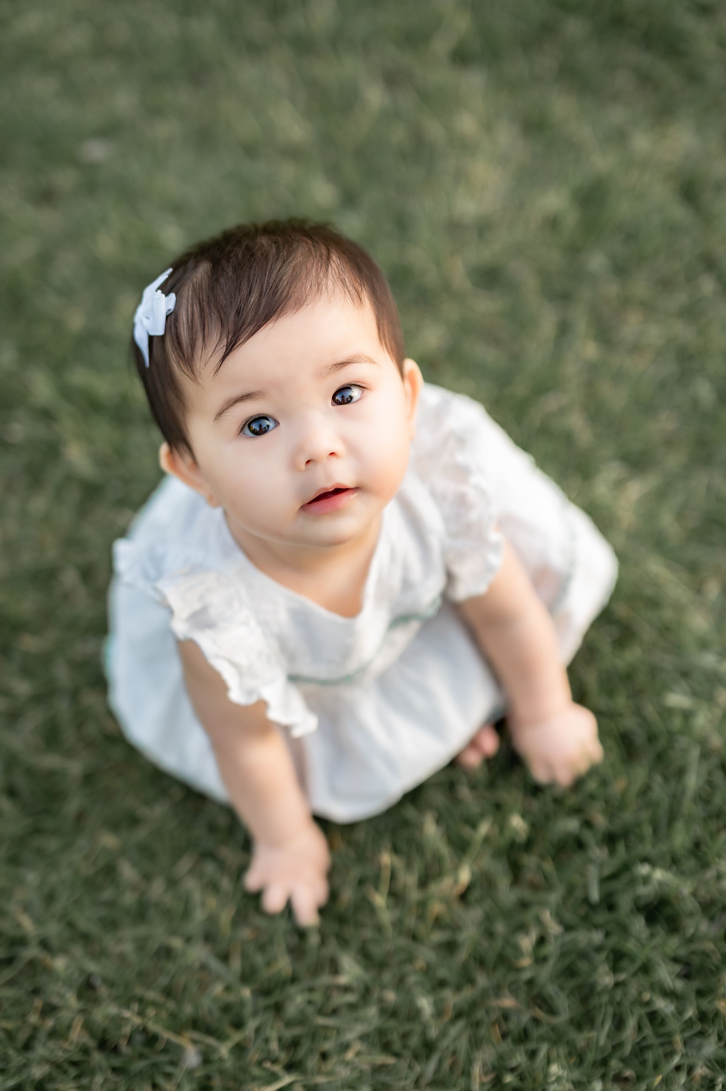 A cute baby girl in a white lace dress and bow sits on green grass, looking up with big brown eyes.