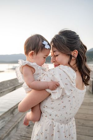 A mother tenderly touches foreheads with her baby girl on a wooden dock at sunset, both wearing white dresses.