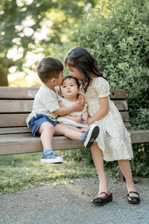 Three young siblings sit together on a wooden park bench, sharing a sweet moment outdoors.