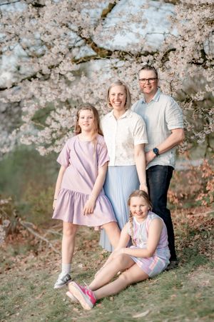Family of four posing under blooming cherry blossom tree, two girls in pink dresses with smiling parents in spring.