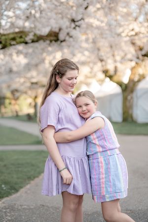 Two girls in pastel dresses embrace under blooming cherry blossom trees in a park during spring.