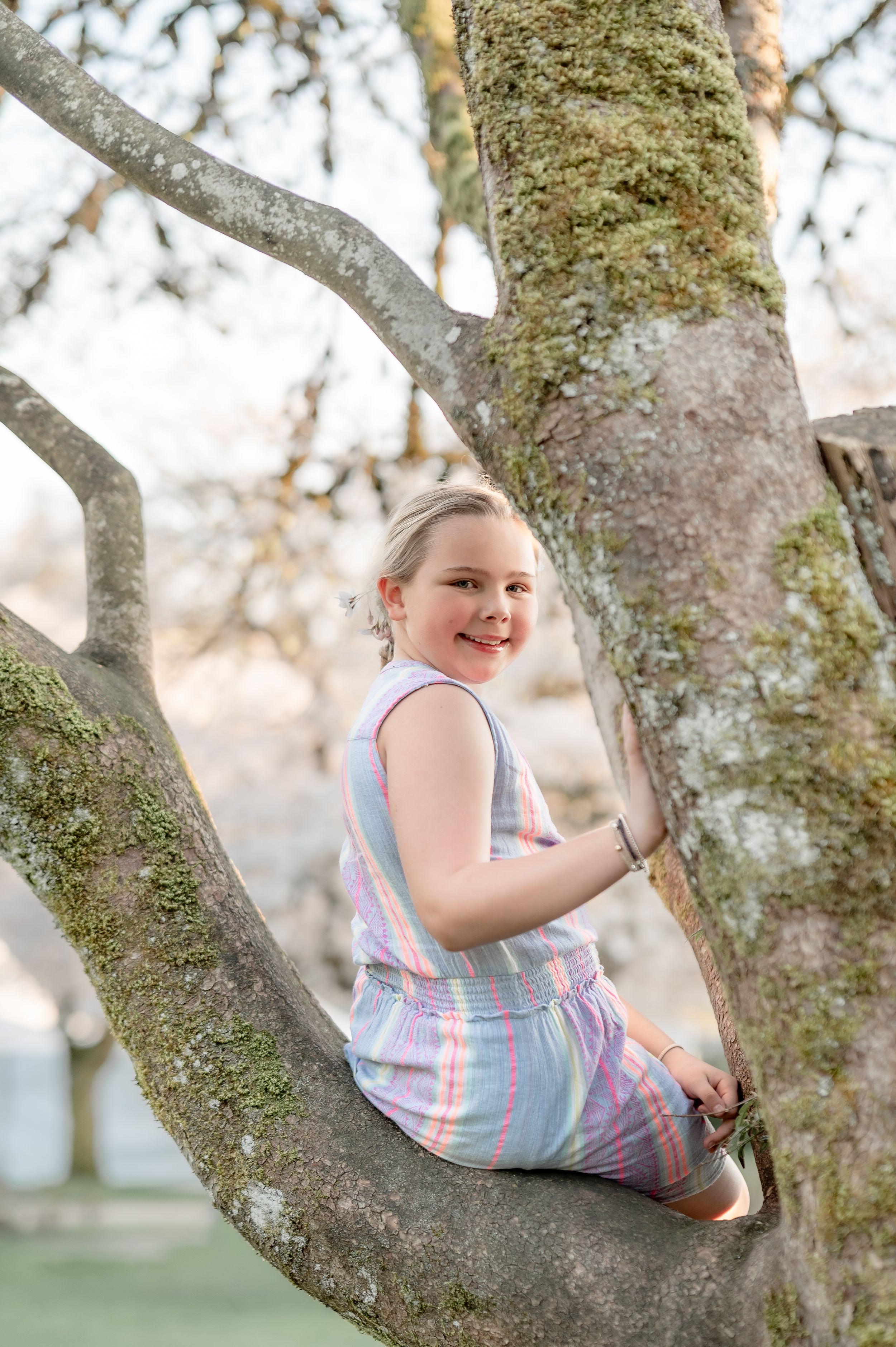 Young girl in pastel romper smiling while sitting in the branches of a moss-covered tree outdoors.
