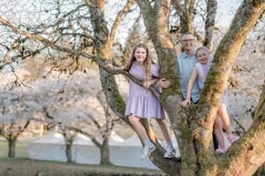 A grandfather poses with two granddaughters in a blooming cherry blossom tree during a spring family photo session.