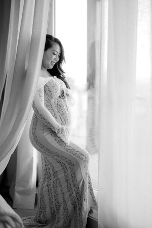 Artistic black and white maternity photos showing a woman in a lace dress posing elegantly by sheer curtains.