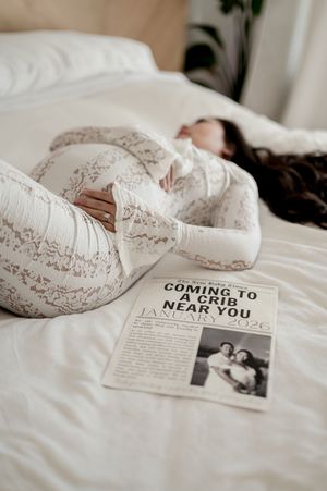 A pregnancy announcement newspaper clipping lies on a white bed next to someone resting in a lace maternity dress.