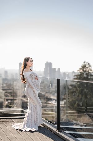 A person in a flowing white lace dress poses elegantly on a rooftop terrace with a cityscape backdrop in soft sunlight.