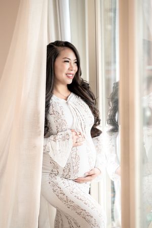 A woman in a white lace dress poses elegantly by sunlit windows in a dreamy maternity photoshoot.