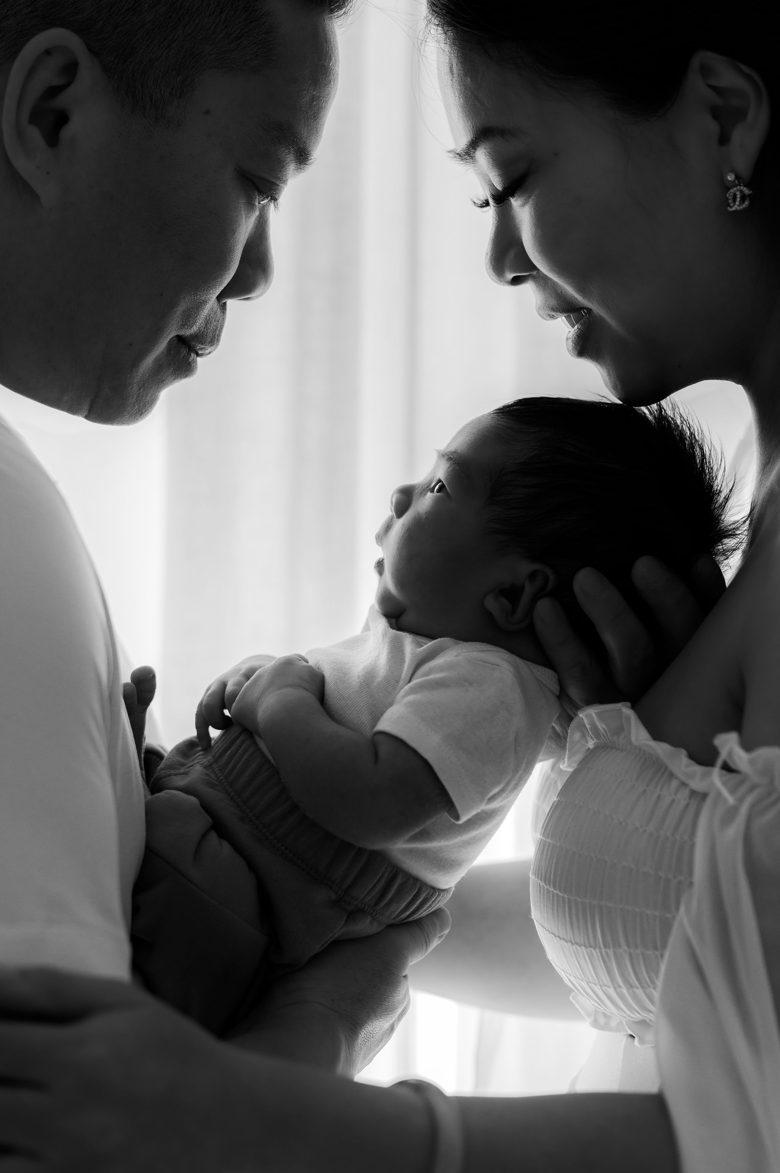Black and white silhouettes show tender moments between parent and newborn in a series of intimate portraits by window light.
