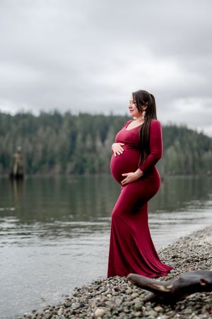 Expectant mother in burgundy dress poses gracefully by a serene lake with forested hills in the background.