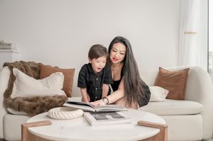 Two people sit together on a beige couch looking at a book on a coffee table in a bright living room.
