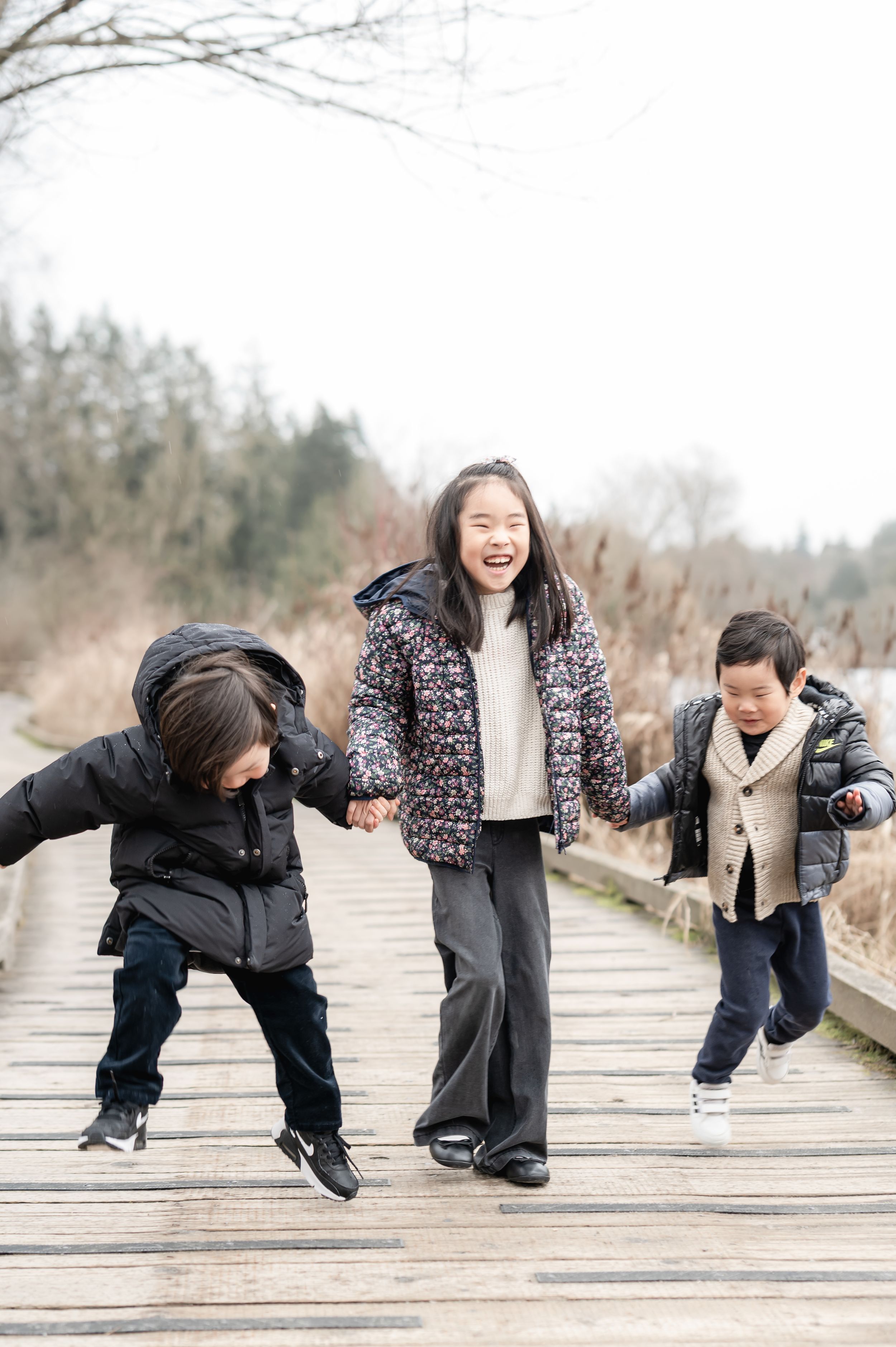 Three children in winter coats playing and laughing together on a wooden boardwalk path on a cloudy day.