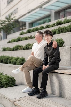Two people sit together on concrete steps outside a modern building with a glass canopy and landscaped greenery.