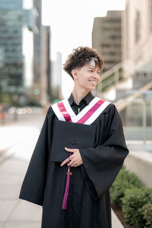 A graduate in black and pink academic regalia smiles while posing outdoors near a building.