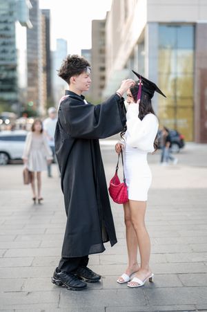 A graduate in black cap and gown helps adjust regalia on someone wearing a white dress during an outdoor ceremony.